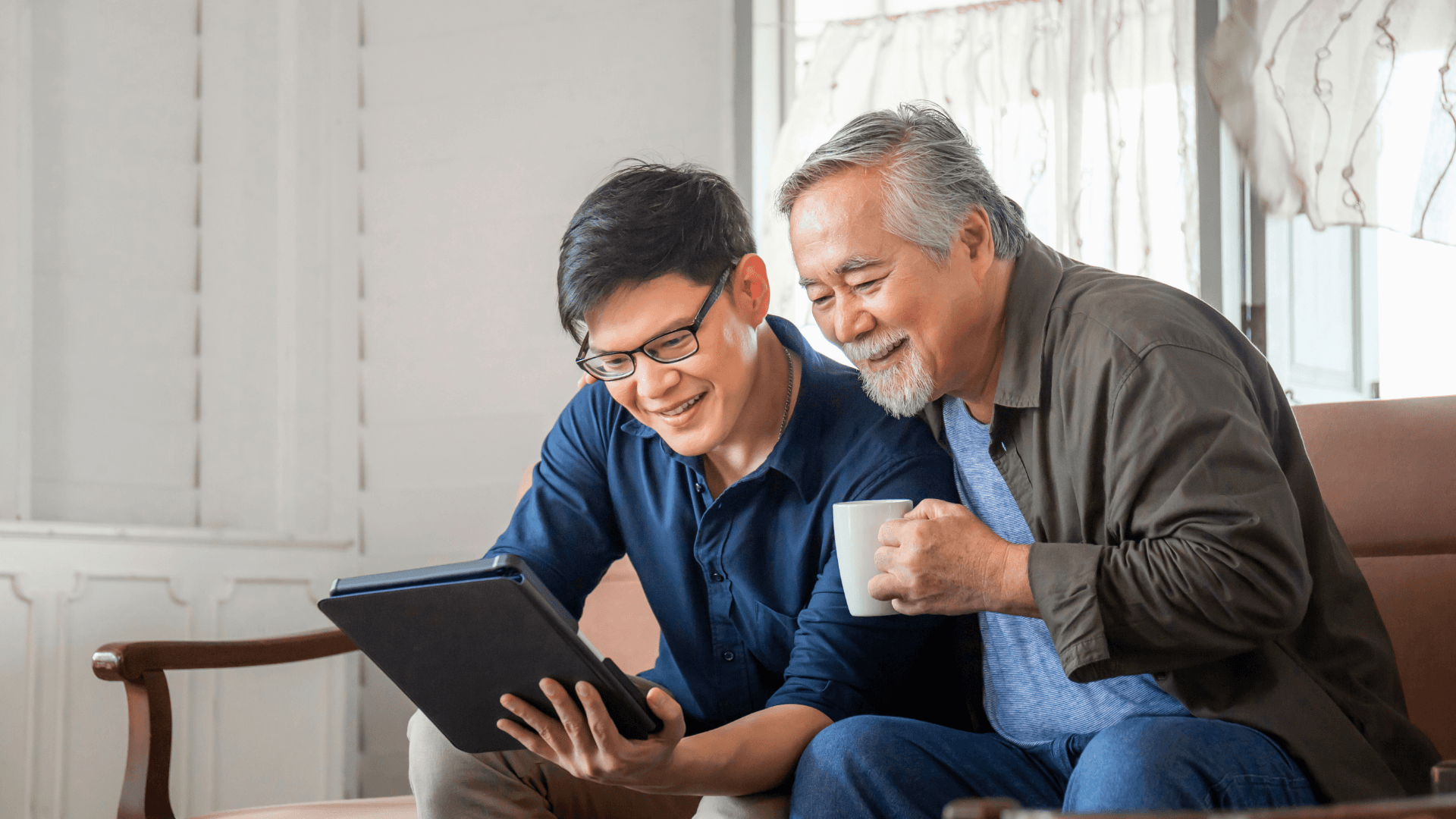 Image of a an elderly male with a cup of coffee with a younger male on a tablet.